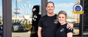 Two smiling staff members in black t-shirts stand outside Joy Bar Coffee Co, promoting a welcoming atmosphere. A certification badge is visible.