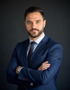 A professional headshot of Mustafa A. Nejem wearing a navy suit and tie, arms crossed, standing confidently against a dark background. He is an award-winning maritime thought leader and author.