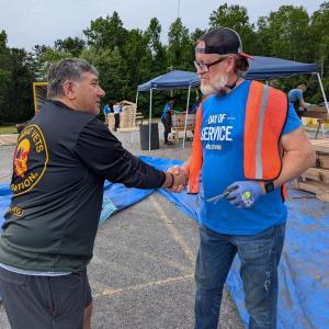 Two veteran volunteers shake hands at a Sleep in Heavenly Peace bed build event. On the left, a man wearing a black VFW (Veterans of Foreign Wars) jacket with patches greets a volunteer on the right wearing a blue 'Day of Service' t-shirt with an orange s