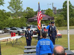 An honor guard ceremony at the Sleep in Heavenly Peace bed build event features three uniformed military personnel presenting colors with an American flag. In the foreground, a volunteer wearing a blue Sleep in Heavenly Peace t-shirt with the organization