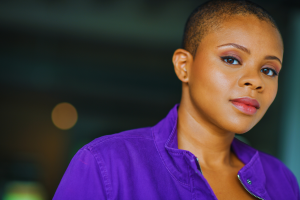 African-American woman with buzz cut, almond eyes and pink full lips seen from the shoulders up, wearing a purple jacket with blurred background- actor Mia Bible promoting her appearance at the 2025 International Black Film Festival.