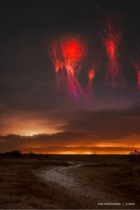 Red bursts of light, known as sprites, illuminate the sky above a field intersected by a dirt path and distance trees.
