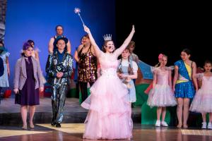 A group of young performers on stage during a theatrical production, with the central actor dressed as Glinda the Good Witch in a pink ball gown and silver crown, holding a sparkling wand overhead and smiling broadly. Other cast members in colorful costum