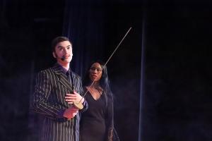 Two actors perform on stage under dramatic lighting. The actor in the foreground, dressed in a dark pinstripe suit with a purple shirt and tie, holds a fencing sword and wears a headset microphone. Behind him, a woman in a long black dress with lace sleev