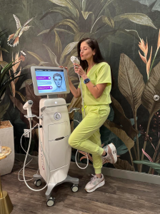 Khush Thanki of Bodyloom Wellness playfully posing with the handheld applicator of a Candela Matrix Pro device, standing in front of a tropical wallpaper backdrop featuring birds and leaves.