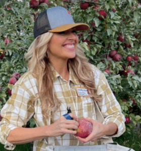 photo of Annessa Chumbley with a SugarBee® hat and picking bag on, while smiling and holding a SugarBee® apple