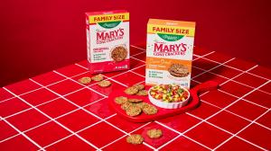 Two boxes of Mary’s Gone Crackers, with crackers scattered on a red tiled surface. A bowl of vegetable dip sits on a red cutting board, surrounded by crackers.