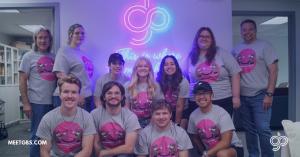 Group photo of the GBS Worldwide team wearing grey 'GBS Summer Camp 2025' shirts with a cartoon llama graphic. The team is smiling and gathered in front of a glowing neon sign that says ‘This is where the magic happens,’ under the GBS logo.