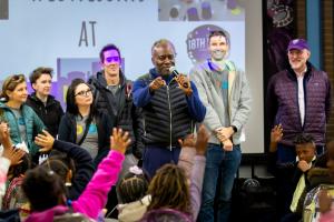 Courtney Williams speaking into a microphone at a KIPP DC event, surrounded by educators and students raising their hands.