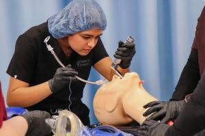 Student wearing scrubs and a surgical cap practices intubation on a medical training manikin during simulation lab