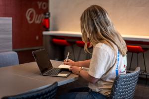 A female student sitting at a table writes in a notebook while looking at an open laptop. The beginning of college life can bring significant mental health challenges for young adults.