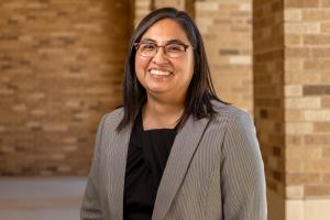 headshot photo of Nancy Treviño, Ph.D., wearing glasses, a black blouse and a gray blazer, standing in a tan brick-walled hallway