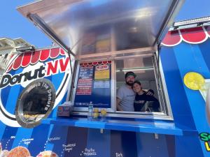 A photo of a husband and wife standing in a food trailer looking at the camera through the window of the DonutNV trailer