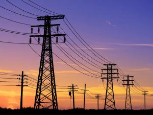 Silhouettes of multiple power line towers and cables against a colorful sunset sky with orange, yellow, and purple hues.