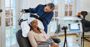 Young woman sitting in chair, getting TMS coil adjusted over her head by healthcare provider, in preparation for depression treatment