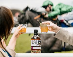 A bottle of Never Say Die Bourbon at Epsom Derby with a racehorse racing past in background