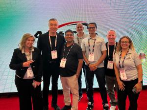 A group of seven AntGen employees stand smiling in front of a digital display with the AntGen logo, wearing name badges. The atmosphere is professional and friendly.