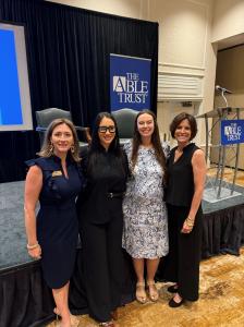 Group photo showing four women standing in front of a stage, dressed in business attire.