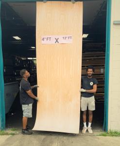 Two men hold a large 4-foot by 12-foot wood veneer sheet outside a warehouse doorway. The sheet has a visible wood grain pattern and a pink sign on top that reads “4’ FT x 12’ FT.” One man stands on each side, supporting the veneer as they carry it.