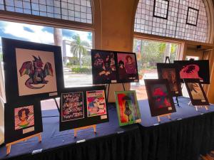 A young LGBTQ+ artist smiles in front of a colorful display of framed drawings, standing beside her own artwork at a community event.