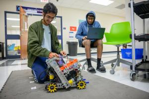 Two students at GSSM work on a robotics project. One student adjusts the robot’s components on the floor while another sits nearby with a laptop, programming.