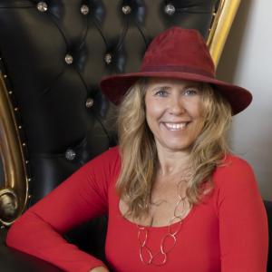 Rachel Anne, founder of Menopause Education Center, smiling while wearing a red hat and red top with gold chain necklace, seated against an elegant black tufted backdrop