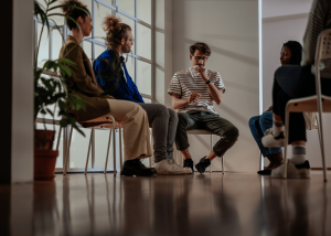 Four diverse adults sitting in a circle on chairs in a bright, modern therapy room with large windows and plants, engaged in what appears to be a group therapy session.