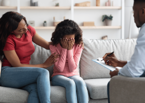 A family therapy session showing a mother comforting her young daughter who is covering her face with her hands, while a therapist takes notes in the background, illustrating integrated family mental health care.