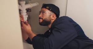 Plumber inspecting kitchen pipes with safety gear before repairing a leak under the sink