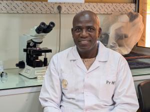 Picture of a sitting man in a lab coat looking at the camera with a microscope behind him