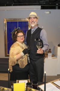 Two people attending the Bay Atlantic Symphony's Roaring 20s Reprise Gala. A man is wearing a suit and hat while holding a glass of wine. The woman next to him is wearing a feathered headband and shimmering shawl. They are smiling together.