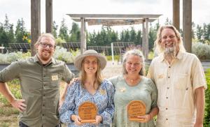 Left to right: Joey Schneiderhan (MRH Sustainability Specialist), Cameron Stearns (MRH CEO), Elise and Jeff Higley (Owners of Oshala Farm). Image feature all four people smiling and holding up two awards from the Oregon Organic Coalition.