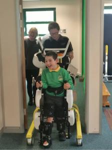 Pediatric patient using a RoboCT gait-training exoskeleton at Morrello Clinic, smiling, with supportive parents nearby and a therapist supervising the session.