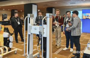 Two smiling participants wearing RoboCT’s lower-limb exoskeleton take supported steps while therapists guide and monitor posture and cadence.