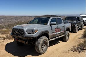 A gray Toyota Tacoma equipped with Ride Shocks suspension tackles the rugged trails of Johnson Valley, with other off-road rigs following behind under a clear desert sky.