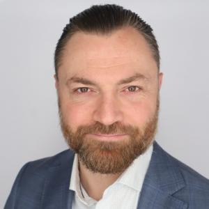 Professional headshot of a man with neatly groomed beard and slicked-back hair, wearing a blue suit jacket over a white patterned shirt, against a light gray background.