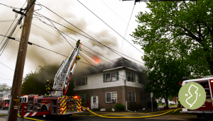 Fire trucks and firefighters on a ladder hose fight heavy smoke from the roof of a two-story NJ home after a heating system fire, stressing the need for heating repair in North NJ.