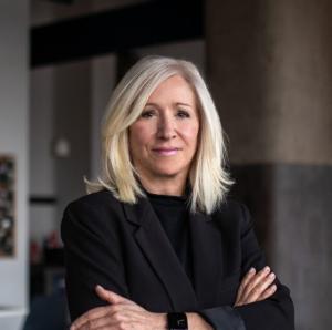 Professional headshot of a confident middle-aged woman with blonde hair, wearing a black blazer, standing with arms crossed in a modern office setting.