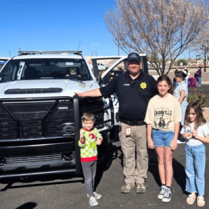 A law enforcement officer stands next to a police truck, posing with three children in a sunny outdoor setting.