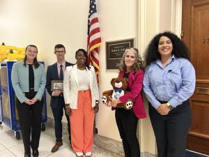 Five people stand smiling outside an office door beneath an American flag. One holds a teddy bear.