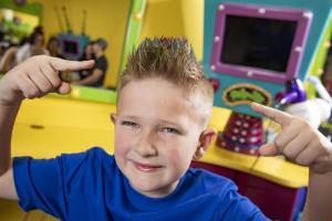 A boy in a blue shirt with blonde hair, smiling in a salon chair and pointing at his hair.