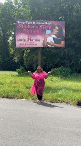 Amber Gilmore wearing a pink dress, smiling with arms open, standing beneath a Creative Nurturing billboard that reads “Amber’s Here Tonight” with the hotline number 844-8-NURTURE