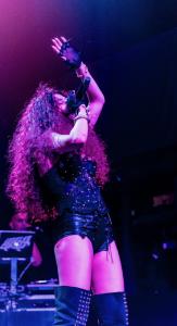 Farrah Mechael performs live in a studded black corset and leather lace-up shorts, paired with tall black boots. Her voluminous curly hair cascades down as she sings into the microphone under purple and blue stage lighting, one arm raised with dramatic in