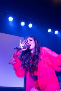 Farrah Mechael on stage in a sheer red feather-trimmed robe layered over her bold stage outfit. With long flowing hair and a glittering choker, she holds the microphone close and gestures expressively with her other hand under bright blue stage lighting.