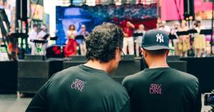 Back view of two SPLEX One team members in branded shirts, standing in front of a stage and looking into a camera filming the performers.