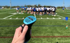 A hand holding the Kühler palm cooling device on the sidelines of a football practice. In the background, a team of athletes and coaches huddles on a turf field under a blue sky.
