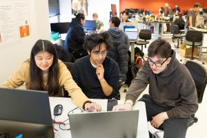 Three students collaborate at a table, working on laptops in a bustling lab. One student gestures toward a screen while the others concentrate. The background shows a busy environment with other students and staff engaged in various technical activities.