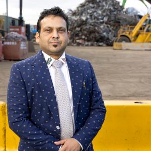 A man in a blue patterned suit standing in front of a scrap metal yard with machinery in the background.