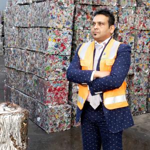 A man in a patterned suit and orange safety vest standing with folded arms in front of large stacks of compacted metal cans and scrap.