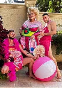Drag queen Buff Faye poses poolside in a colorful costume with two shirtless men wearing pink “EAT IT UP” aprons and accessories, celebrating a playful bachelorette cookie decorating party vibe with a large pink beach ball.